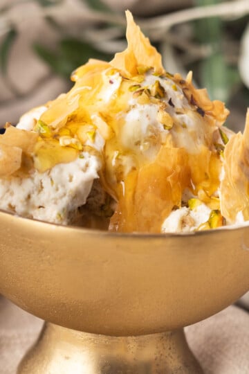 Gold bowl with no-churn baklava ice cream, honey drizzle, pistachios, and phyllo pastry shards, set on a rustic background.