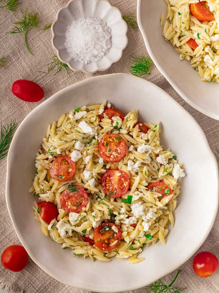 Tomato and feta orzo salad with cherry tomatoes, feta cheese, fresh herbs, and a dish of sea salt on a beige tablecloth.