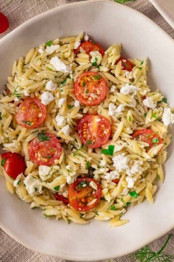 Tomato and feta orzo salad with cherry tomatoes, feta, and herbs in a white bowl on a textured tablecloth.