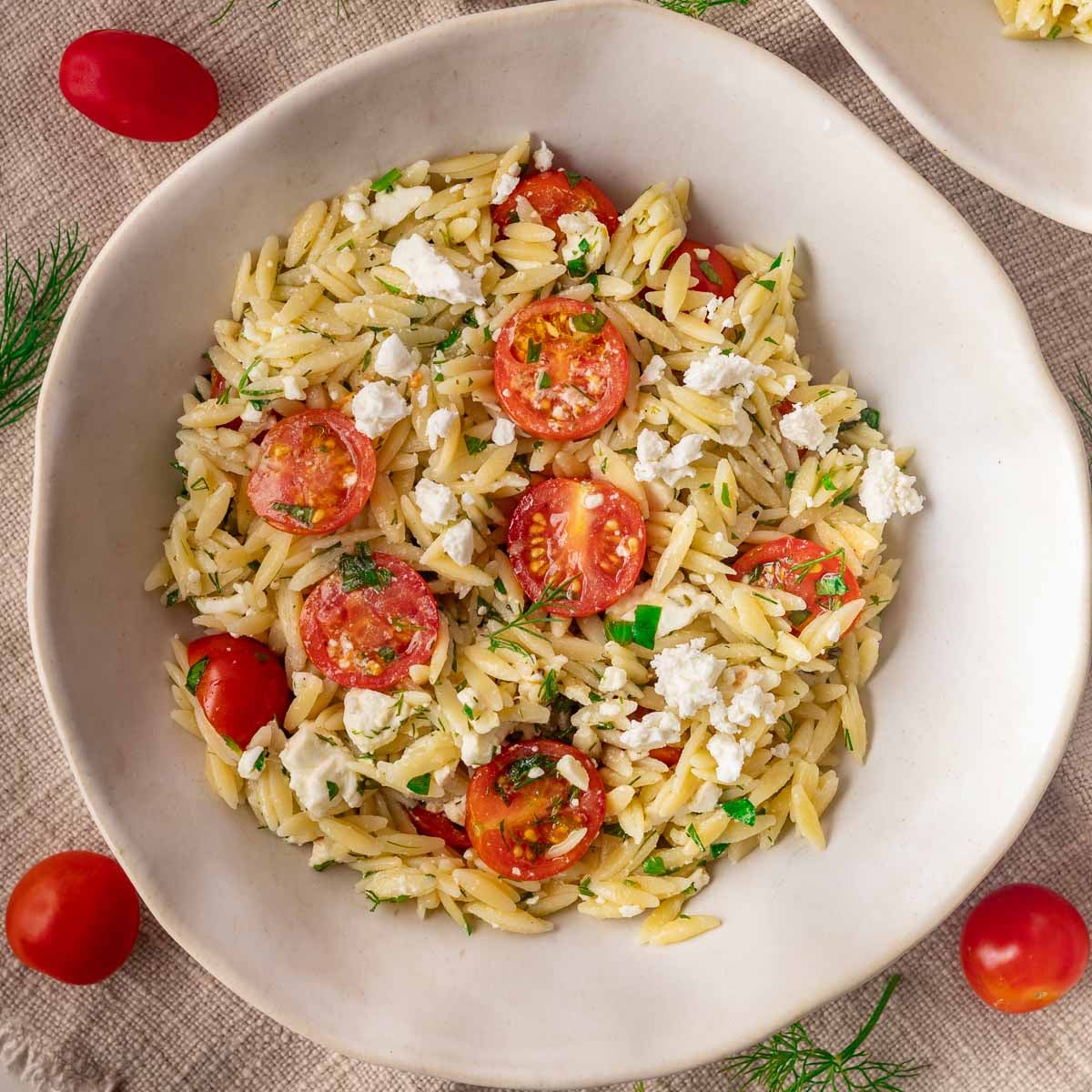 Tomato and feta orzo salad with cherry tomatoes, feta, and herbs in a white bowl on a textured tablecloth.