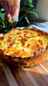 Hand garnishing a quiche with chives and bacon on a wooden board.