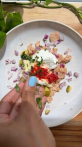Hand adding herbs to tuna salad with diced onions and sauce in a white bowl.