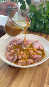 Pouring marinade over raw chicken pieces in a white bowl on wooden table.
