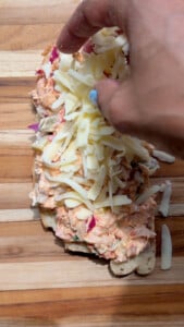 Hand adding grated cheese on open-faced tuna salad sandwich on wooden board.