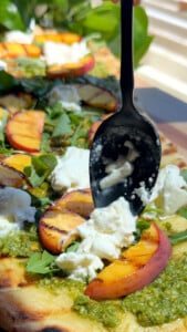 Grilled peach and burrata flatbread with pesto and arugula being prepared with a spoon.
