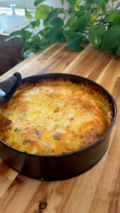 Freshly baked quiche in pan on wooden table with green plants in background.