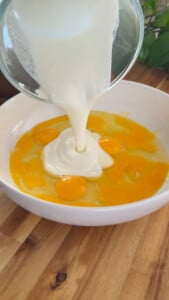 Pouring milk into beaten eggs in a white bowl on a wooden table.
