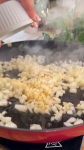 Chopped onions sautéing in a skillet, hand adding more onions from a white bowl.