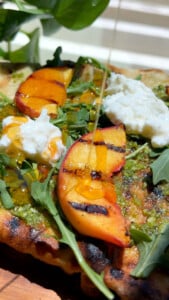 Grilled peach and burrata flatbread with arugula, pesto, and hot honey drizzle in sunlight.