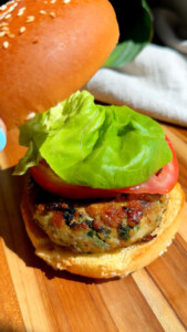 Homemade chicken veggie burger with lettuce and tomato on a sesame seed bun on a wooden surface.