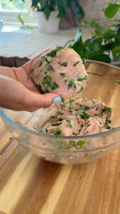 Person forming a chicken burger patty with spinach and herbs in a glass bowl on a wooden countertop.
