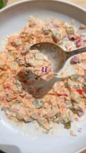 Spoon scooping creamy tuna salad with diced pickles and onions in a white bowl.