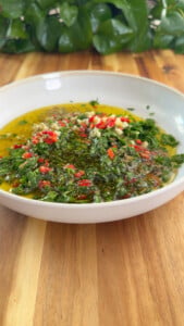 Chimichurri sauce with chopped herbs and red peppers in a white bowl on wooden table.