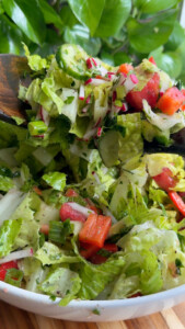 Fresh salad with lettuce, cucumber, tomatoes, radishes, and peppers in a white bowl with a wooden spoon.