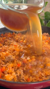 Pouring broth into a pan of rice, vegetables, and meat.