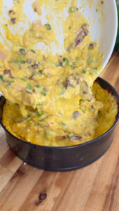 Pouring cheesy mixture with green onions and bacon into a baking pan on a wooden surface.