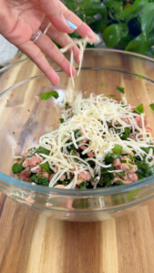 Hand adding cheese to chicken mix with herbs in glass bowl on wooden surface.