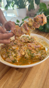 Person skewers marinated chicken with herbs in a bowl, on a wooden table.