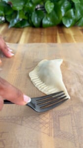 Sealing empanada dough with a fork on parchment paper with green leaves in the background.