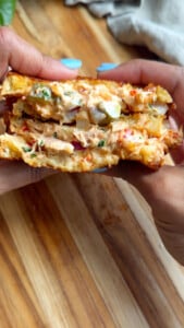 Grilled tuna sandwich with vegetables, and creamy filling, held in hands over wooden surface.