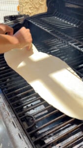 Hands placing flat dough on a barbecue grill.
