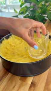Pressing mashed potatoes into a cake pan using a glass on a wooden countertop with a plant in the background.