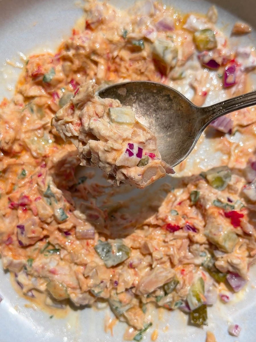 Close-up of a spoon scooping colorful tuna salad with pickles and onions in a white bowl.