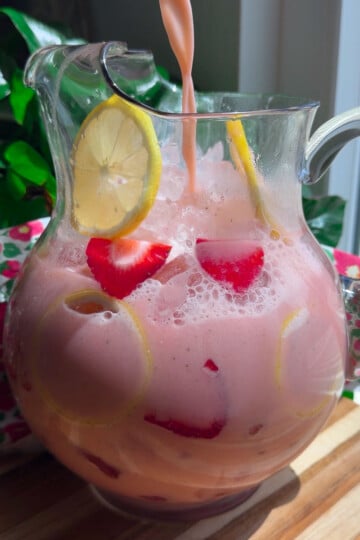 Refreshing pink lemonade with sliced strawberries and lemons in a clear jug, set on a wooden table.