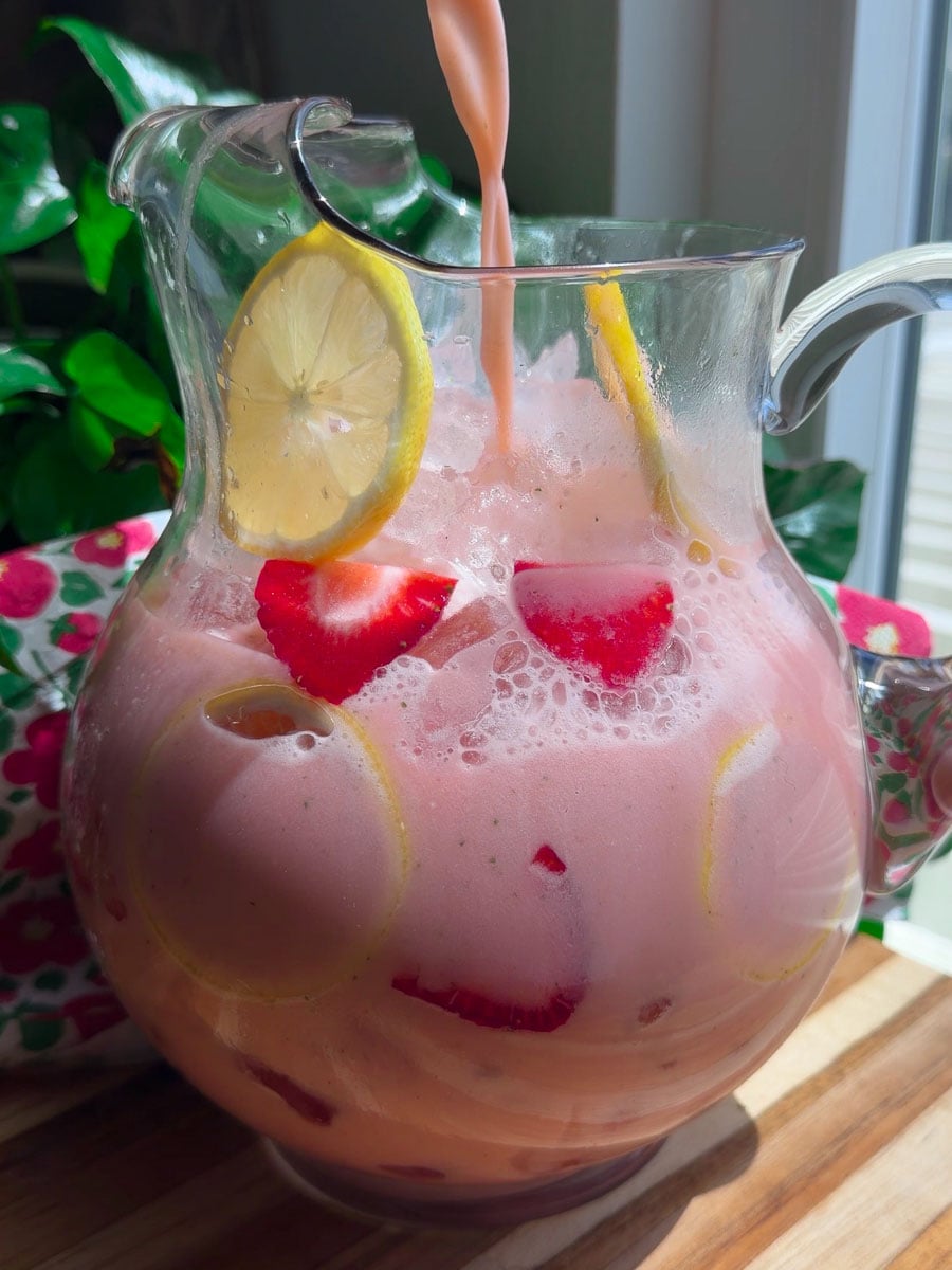Refreshing pink lemonade with sliced strawberries and lemons in a clear jug, set on a wooden table.