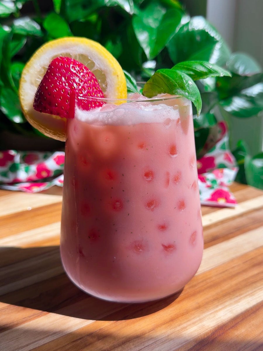 Refreshing strawberry lemonade with ice, garnished with a lemon slice and basil, on a wooden table.
