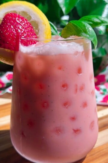 Pink strawberry lemonade with ice, garnished with lemon slice, strawberry, and basil leaves on wooden table.