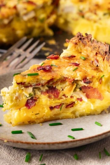 Slice of Potato Crust Quiche with bacon and chives on a plate, garnished with fresh herbs.