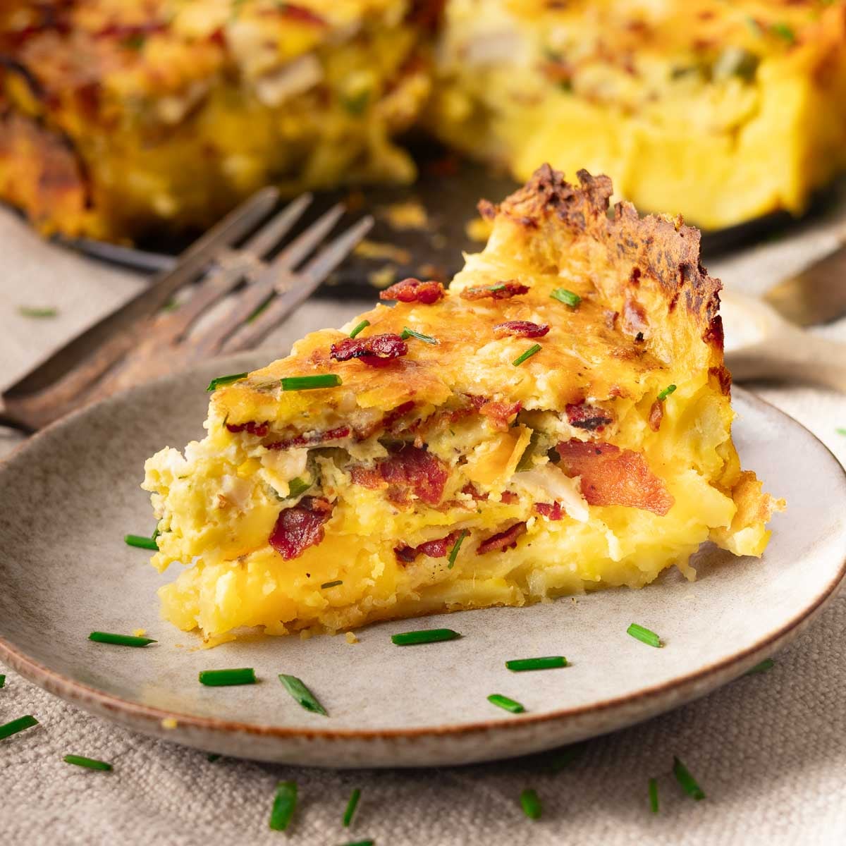 Slice of Potato Crust Quiche with bacon and chives on a plate, garnished with fresh herbs.