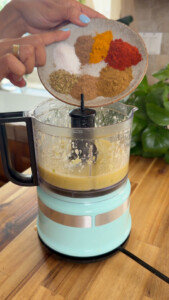 Adding spices to a light blue food processor with sauce on a wooden kitchen counter.