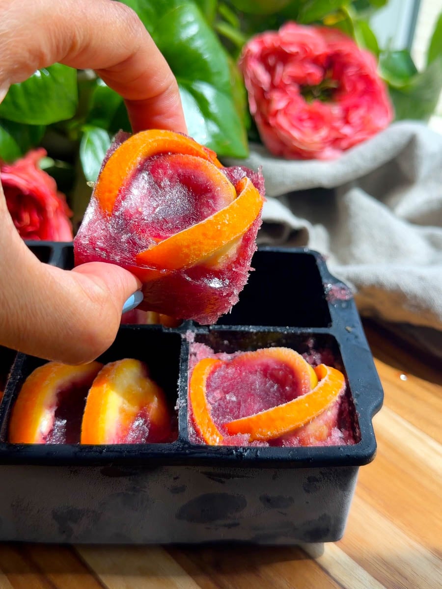 Tinto de verano ice cube in a hand above a black tray, with pink roses in the background.