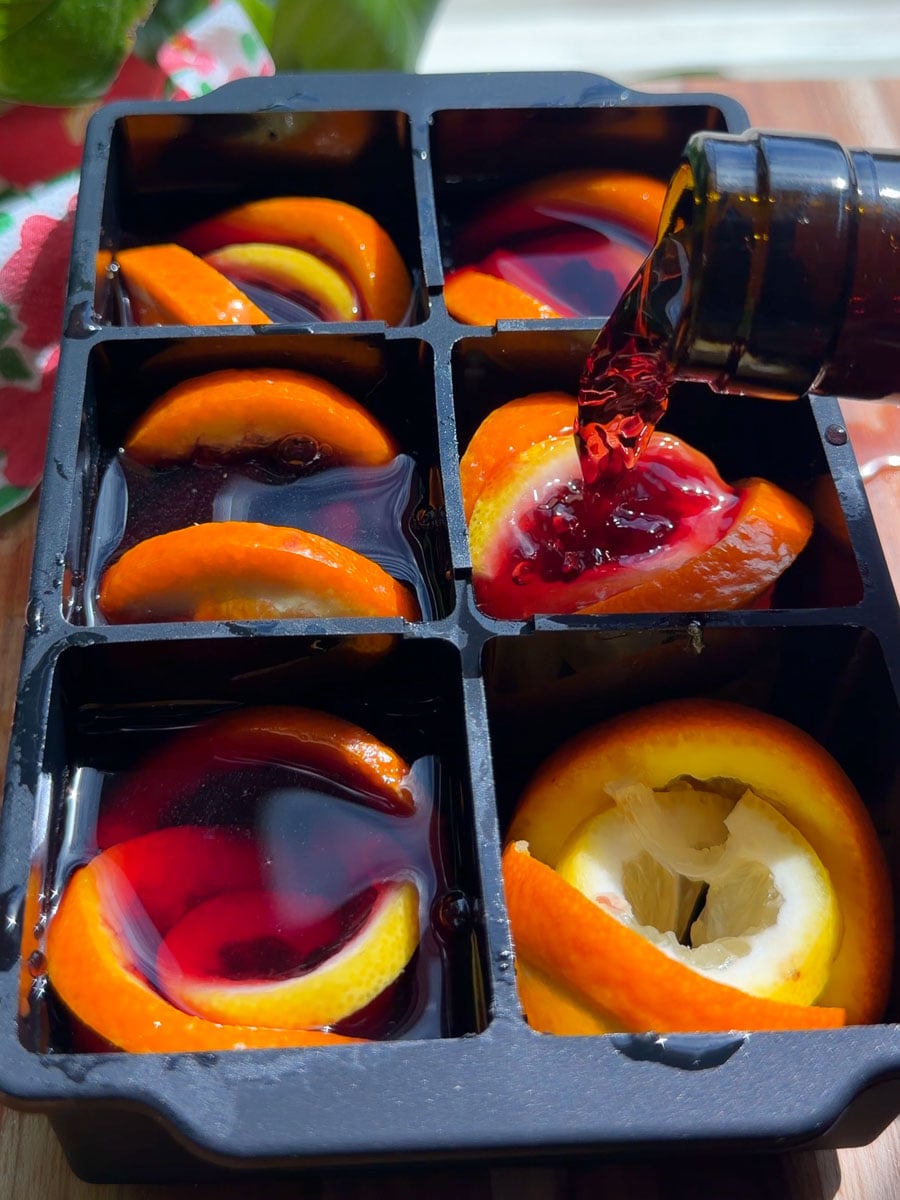 Pouring red liquid into ice cube tray with orange and lemon slices to make Tinto de verano cube.