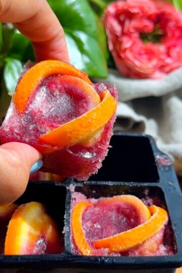 Hand holding tinto de verano ice cube with orange peels over mold, with flowers in the background.