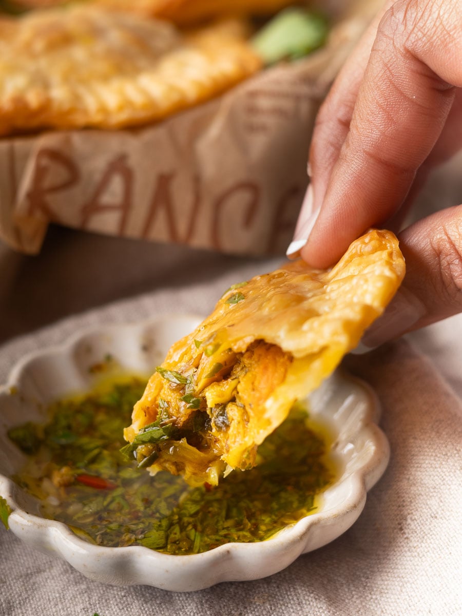 Close-up of a hand dipping a tuna pastel into chimichurri sauce, showcasing its crispy texture and flavorful filling.