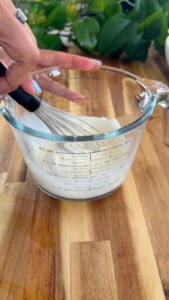 Hand whisking liquid in a glass measuring cup on a wooden countertop.