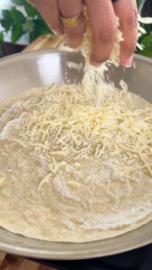 Hand sprinkling cheese on a tortilla in a pan.