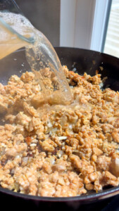 Pouring broth over ground meat sizzling in a pan by the window.