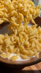 Pouring shell pasta into a wooden bowl filled with creamy yogurt sauce.