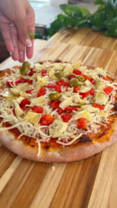 Homemade pizza topped with cheese, red peppers, artichokes, green olives on wooden board.
