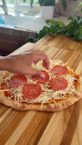 Hand placing pepperoni slices on cheese pizza on wooden countertop.