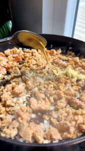 Adding mustard to cooking ground sausage in skillet near window.