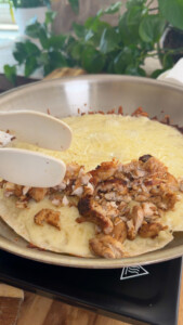 Chicken quesadilla cooking in a pan with cheese and shredded chicken under tongs.