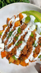 Loaded potato dish with ground meat, avocado slices, drizzled with white sauce, garnished with cilantro.