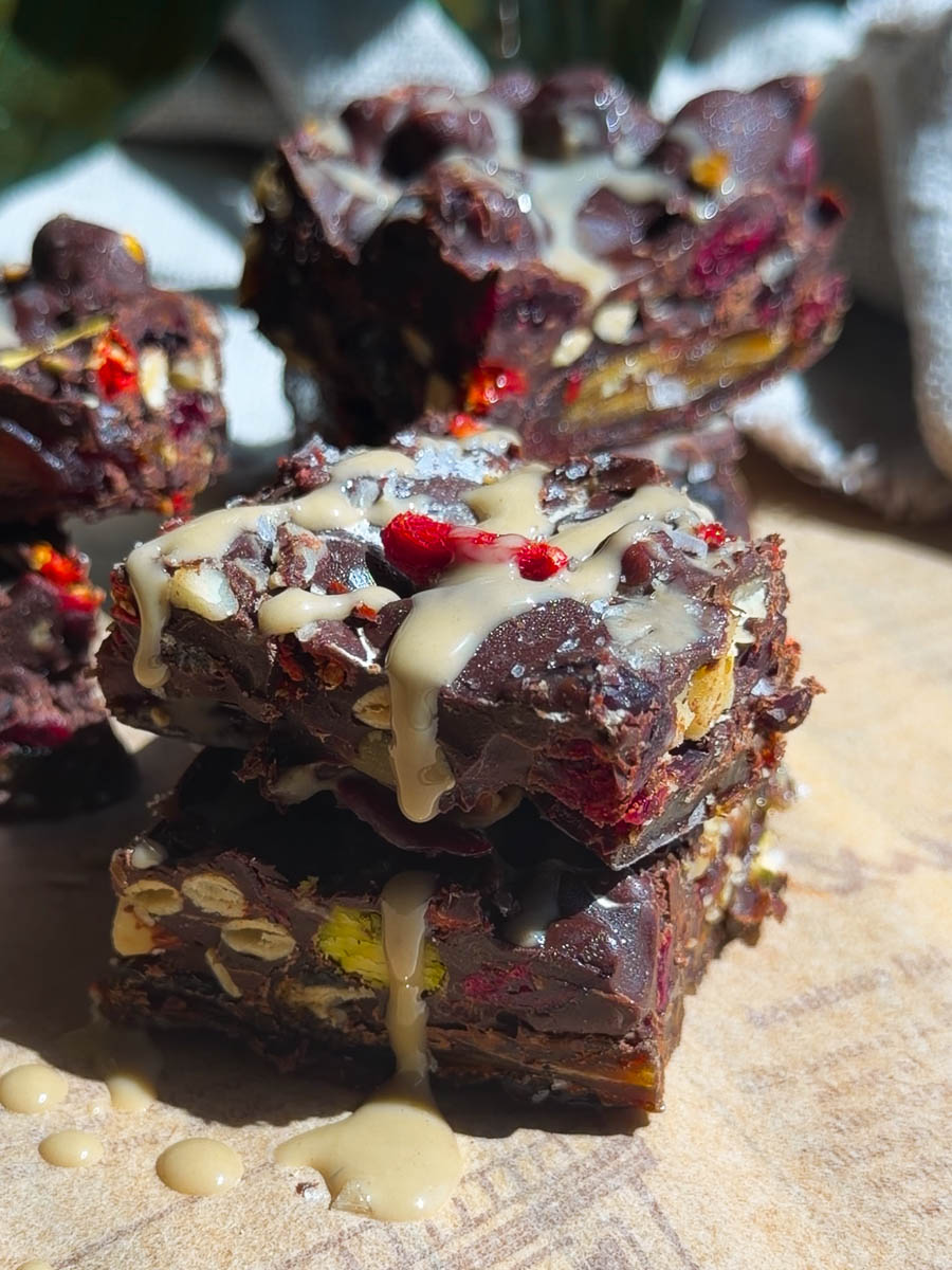 Close-up of stacked chocolate tahini date bark with nuts, berries, and a drizzle of white glaze on top.