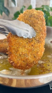 Tongs holding crispy breaded cutlet over frying pan with oil, close-up.