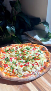 Mediterranean pizza with olives, artichokes, and red peppers on a wooden board near plants.